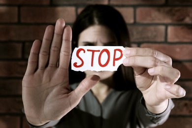 Photo of Domestic violence concept. Woman holding paper with word Stop near brick wall, focus on hands