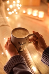 Woman with cup of drink on floor against blurred Christmas lights, closeup