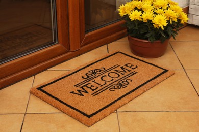 Door mat with word Welcome and beautiful flowers on floor near entrance