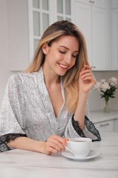 Pretty young woman in beautiful silk robe with cup of coffee at kitchen table