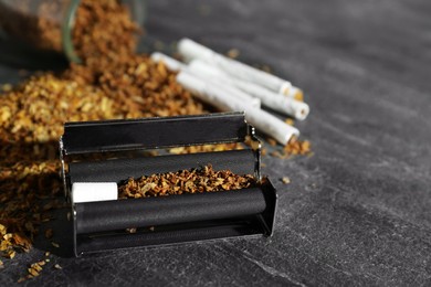 Photo of Roller, tobacco and hand rolled cigarettes on dark grey table, closeup. Space for text