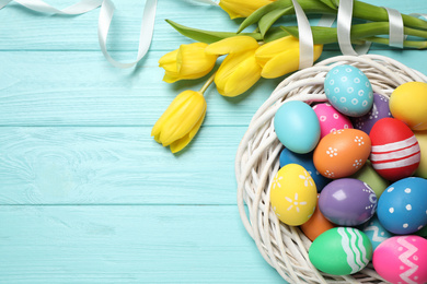 Colorful Easter eggs in decorative nest and tulip flowers on light blue wooden background, flat lay. Space for text
