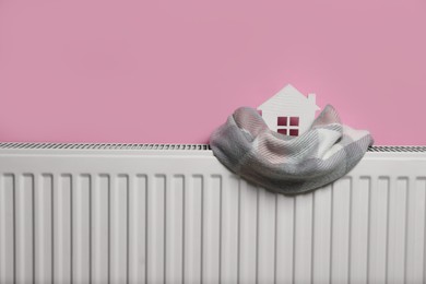 Modern radiator with knitted scarf and wooden house near pink wall indoors, space for text. Winter heating efficiency