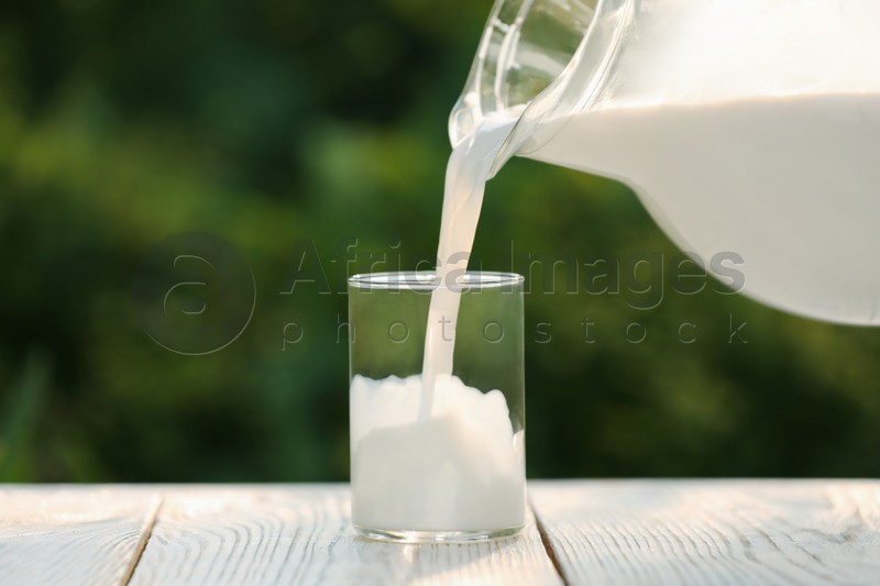 Pouring tasty fresh milk from jug into glass on white wooden table, closeup