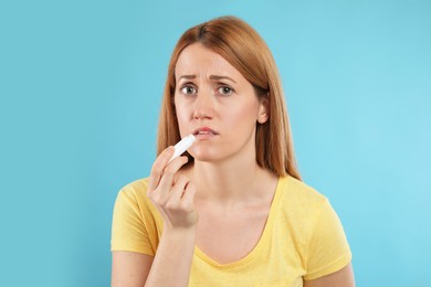 Photo of Upset woman with herpes applying lip balm against light blue background