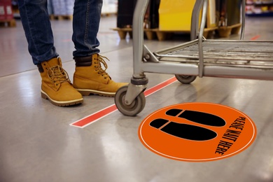 Image of Keep social distance as preventive measure during coronavirus outbreak. Orange warning sign on floor in front of buyer with shopping cart, closeup