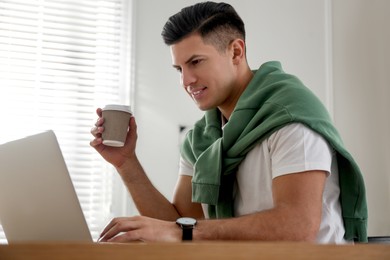 Freelancer with cup of coffee working on laptop at table indoors