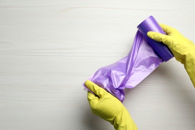 Janitor in rubber gloves holding roll of violet garbage bags over white wooden table, top view. Space for text
