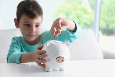 Little boy putting coin into piggy bank at white table indoors