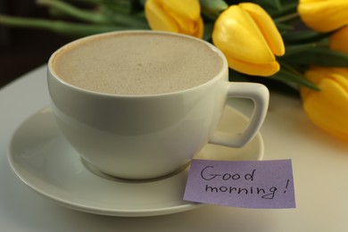 Cup of coffee, flowers and card with phrase GOOD MORNING! on white table, closeup