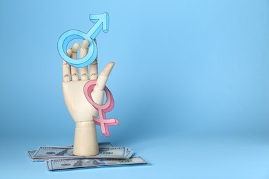 Photo of Gender pay gap. Wooden mannequin hand with symbols and dollar banknotes on light blue background, space for text