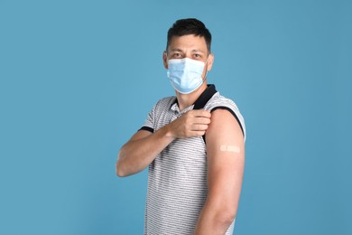 Vaccinated man with protective mask showing medical plaster on his arm against light blue background