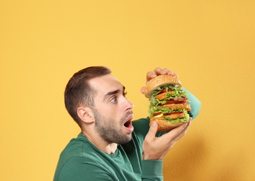 Young hungry man eating huge burger on color background
