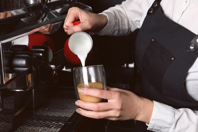 Photo of Barista pouring steamed milk from pitcher into glass cup of fresh aromatic coffee in cafe, closeup