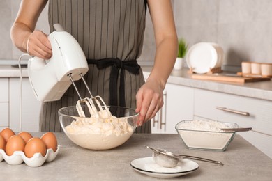 Woman whipping white cream with mixer at light grey table in kitchen, closeup