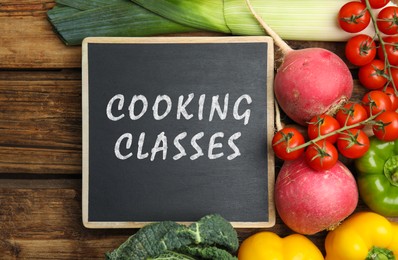 Image of Small chalkboard with inscription Cooking Classes and different fresh vegetables on wooden table, flat lay