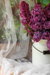 Beautiful lilac flowers in milk can near window indoors, closeup