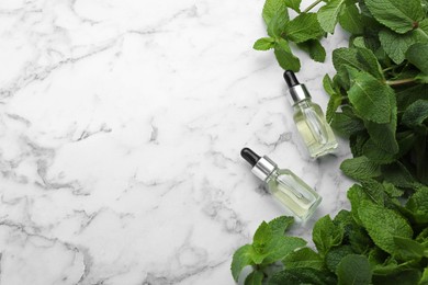 Bottles of mint essential oil and green leaves on white marble table, flat lay. Space for text