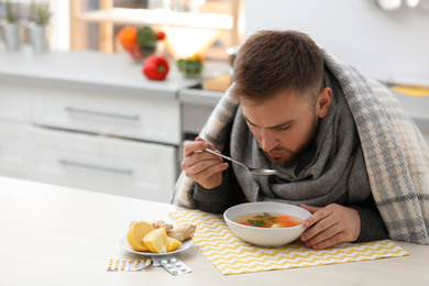 Sick young man eating tasty soup to cure flu at table in kitchen