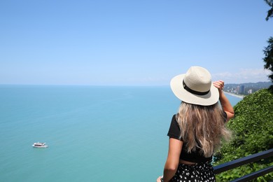 Woman with hat near sea on sunny day, back view. Space for text