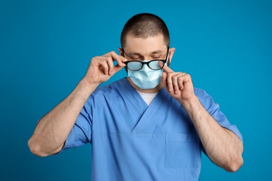Doctor wiping foggy glasses caused by wearing disposable mask on blue background. Protective measure during coronavirus pandemic