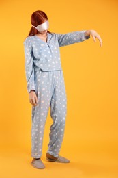 Young woman wearing pajamas, mask and slippers in sleepwalking state on yellow background