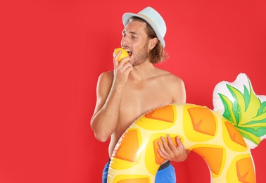 Attractive young man in swimwear with pineapple inflatable ring eating lemon on red background