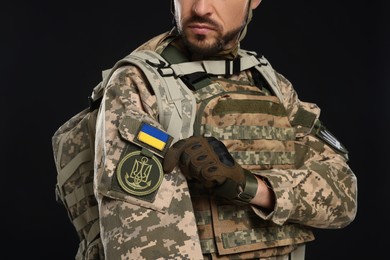 Soldier with Ukrainian flag and trident on military uniform against black background, closeup