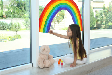Little girl drawing rainbow on window with paints indoors. Stay at home concept
