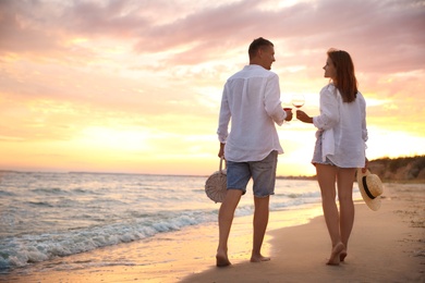Lovely couple with glasses of wine walking on beach at sunset, back view. Space for text