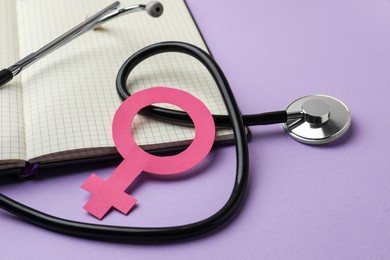 Photo of Female gender sign, open notebook and stethoscope on violet background, closeup. Women's health concept