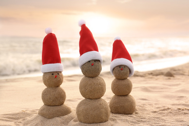 Snowmen made of sand with Santa hats on beach near sea at sunset. Christmas vacation