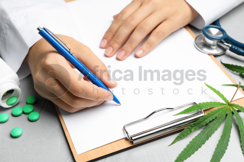 Female doctor with clipboard, pills and hemp leaf at grey table, closeup
