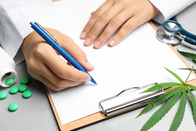 Female doctor with clipboard, pills and hemp leaf at grey table, closeup