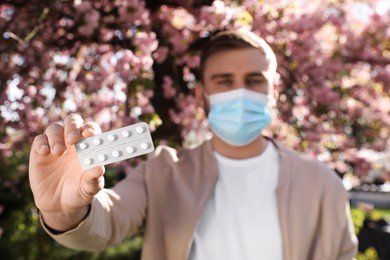 Man with pills and protective mask near blossoming tree outdoors, focus on hand. Seasonal pollen allergy