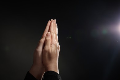 Woman holding hands clasped while praying against light in darkness, closeup. Space for text