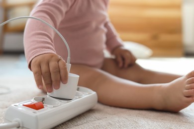 Photo of Cute baby playing with charger and power strip on floor at home, closeup. Dangerous situation