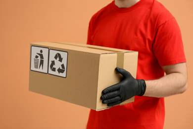 Courier holding cardboard box with different packaging symbols on orange background, closeup. Parcel delivery