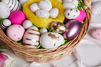 Photo of Wicker basket with festively decorated Easter eggs on white marble table, above view