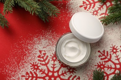 Photo of Winter skin care. Hand cream near snowflake silhouettes made with artificial snow and fir branches on red background, flat lay