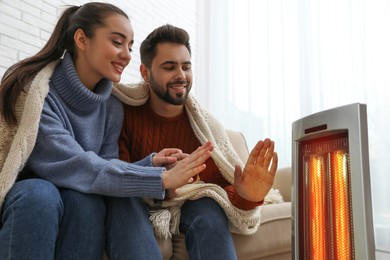 Young couple warming hands near electric heater at home
