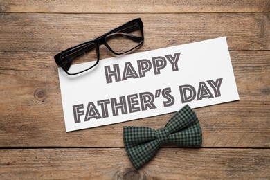 Card with phrase HAPPY FATHER'S DAY, eyeglasses and bow tie on wooden background, flat lay