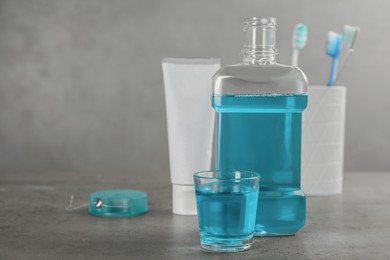 Bottle and glass with mouthwash near other oral hygiene products on grey table, space for text