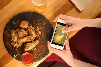 Woman playing game using smartphone at table with tasty BBQ wings, top view