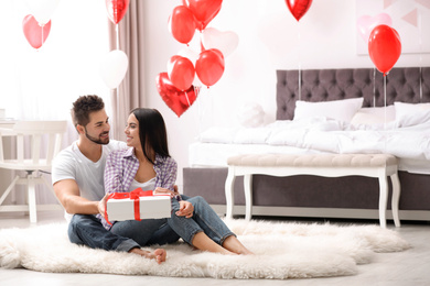 Young man presenting gift to his girlfriend in bedroom decorated with heart shaped balloons. Valentine's day celebration