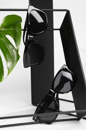 Stylish sunglasses hanging on black shelf against white background