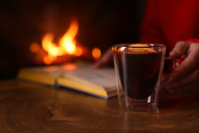 Woman with mulled wine reading book near fireplace indoors, closeup. Winter vacation