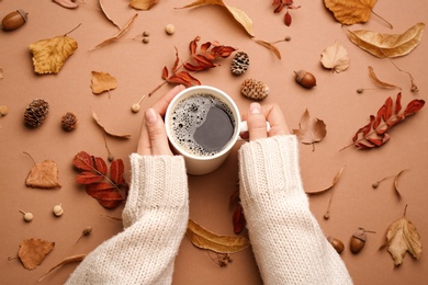Woman with cup of hot drink surrounded by autumn leaves on brown background, flat lay. Cozy atmosphere