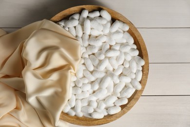 Cocoons with bowl and silk fabric on white wooden table, flat lay