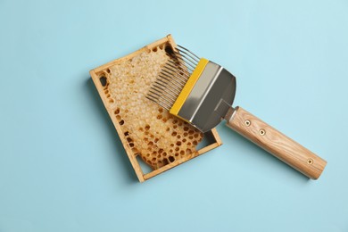 Hive frame with honeycomb and uncapping fork on light blue background, flat lay. Beekeeping